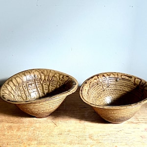 May include: Two handmade ceramic bowls with a textured, organic design. The bowls are a warm, earthy brown color with a unique pattern on the interior and exterior. The bowls are set on a wooden surface.