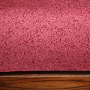 May include: A burgundy paisley patterned table runner. The runner is rectangular with rounded corners and is draped over a wooden surface. The paisley pattern is a darker shade of burgundy.