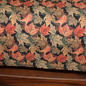 May include: A quilted table runner featuring a repeating pattern of autumn leaves in shades of red, orange, green, and brown against a black background. The leaves are detailed and create a seasonal theme.