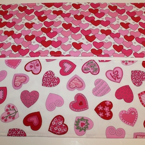 May include: Two white fabric placemats with a pink and white heart pattern. The top placemat has a smaller, more densely packed heart pattern. The bottom placemat has a larger, more spaced out heart pattern with different designs in each heart.