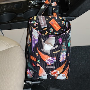 May include: A black fabric car trash bag featuring a repeating pattern of colorful gnomes, candy, and the text "Gnomies" printed on the exterior. The bag has a fabric strap at the top for hanging from a gear shift or other interior vehicle component.