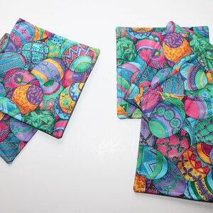 May include: A set of colorful, quilted fabric coasters. The coasters feature a vibrant pattern of Easter eggs in shades of pink, blue, green, and orange. The edges are finished with a dark border. The coasters are square and appear to be made of cotton.