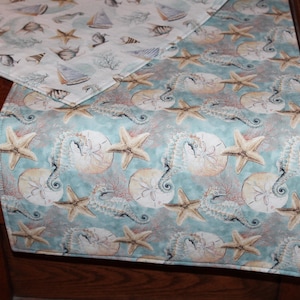 May include: A table runner with a nautical theme. The runner features a repeating pattern of starfish, seahorses, and sand dollars on a teal background. A matching placemat with boats, fish, and shells is visible.