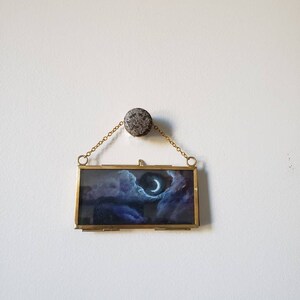 May include: A small, rectangular framed artwork depicting a night sky with a crescent moon peeking through dark clouds. The artwork is encased in a gold-colored metal frame and suspended by a delicate chain from a textured, round, gray stone.