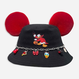 May include: A black bucket hat featuring two large red velvet ears on top. The front displays a graphic of a character dressed as a devil holding a pumpkin. A silver-toned metal chain wraps around the base, adorned with six small hanging charms.