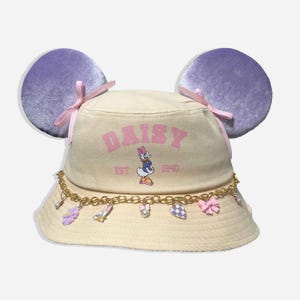 May include: A cream-colored bucket hat with purple mouse ears and pink bows. The hat features the word "DAISY" in pink, along with a Daisy Duck graphic and the text "EST 1940". A gold chain with charms adorns the brim.