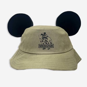 May include: A khaki bucket hat with black Mickey Mouse ears and a black and white graphic of a dinosaur holding a balloon. The graphic also includes the text "WRONGPARK".
