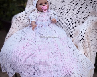 May include: A baby doll wearing a white crocheted christening gown and bonnet, with a pink pacifier. The gown has a long skirt and intricate lace details. The doll is seated on a crocheted blanket.