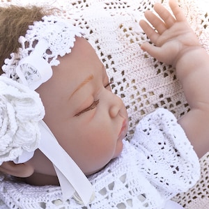 Puede incluir: Una diadema blanca de ganchillo con una flor y una cinta de satén blanca, puesta por una muñeca bebé dormida.