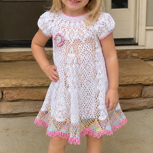 May include: A white crochet dress featuring short puff sleeves, a round neckline, and a floral pattern. The dress is accented with a pastel-colored crochet flower on the chest and a matching pastel-colored crochet trim along the hem and headband.