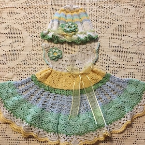 Pode incluir: Um vestido de bebé de crochê com um esquema de cores amarelo, verde e azul. O vestido tem um chapéu de crochê a condizer com um padrão de riscas amarelo, verde e azul.