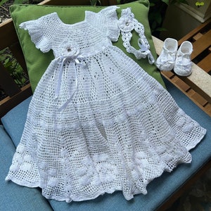 Puede incluir: Vestido de bebé blanco de ganchillo con una cinta de satén y un adorno de flor. El vestido tiene un dobladillo festoneado y se combina con una diadema y zapatos de ganchillo a juego.