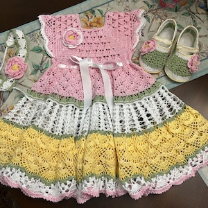 Op de afbeelding: Een handgemaakte gehaakte jurk met een roze lijfje met bloemaccent, een wit satijnen lint en een rok met meerdere lagen in roze, wit, groen en geel. De set bevat een bijpassende gehaakte hoofdband en een paar groen-witte schoentjes.