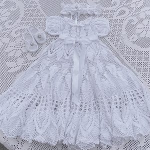 Puede incluir: Un vestido de bautizo blanco de ganchillo con sandalias y diadema a juego. El vestido presenta un diseño calado detallado, mangas cortas y una faja de cinta de raso. Las sandalias tienen un pequeño adorno decorativo.