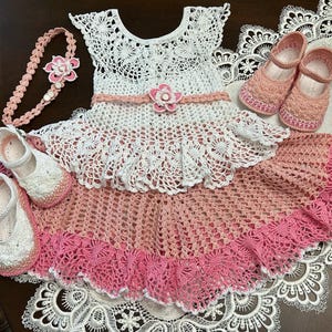 May include: A white and peach crochet dress with a flared skirt, featuring a pink flower accent at the waist. The set includes a matching peach crochet headband with a flower, a pair of white crochet shoes, and a pair of peach crochet shoes.