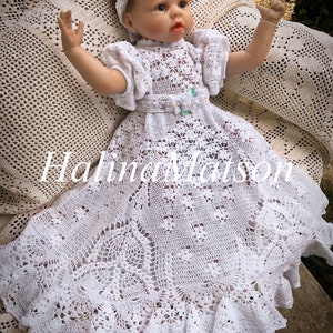 Puede incluir: Un vestido de bebé blanco de ganchillo con una diadema floral. El vestido tiene un dobladillo festoneado y un delicado patrón de encaje.