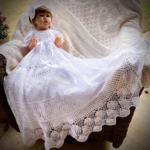 Puede incluir: Un vestido de bebé de crochet blanco con una cinta de satén y un borde de encaje. El vestido se muestra sobre una manta de crochet blanco.