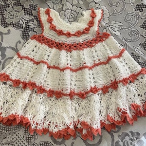 May include: A white crocheted baby dress with a pink and white trim. The dress has a ruffled skirt and a sleeveless bodice.