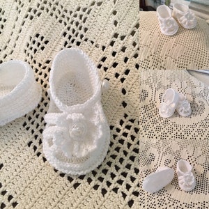 Op de afbeelding: Witte gehaakte babysandalen met een bloemenversiering en parel accenten. De sandalen liggen op een wit gehaakt kantje.