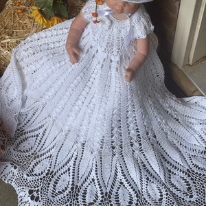May include: A white crocheted baby gown with a delicate lace pattern. The gown has a round, scalloped hem and a small, white crocheted hat with a flower embellishment.