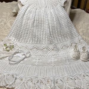 Puede incluir: Un vestido de bebé blanco de ganchillo con un borde de encaje y un gorro a juego. El vestido tiene un corpiño fruncido y una falda completa. El gorro tiene un delicado borde de encaje.
