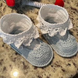 May include: A pair of baby booties in light blue and white crochet. The booties have a white lace trim and a button detail.