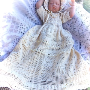 Puede incluir: Un vestido de bebé de ganchillo blanco con un gorro a juego. El vestido tiene un delicado encaje y un borde festoneado. El bebé duerme plácidamente en el vestido.