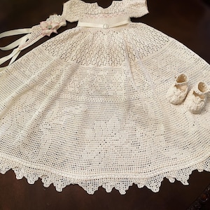 May include: An off-white, crocheted baby dress with matching booties and a headband. The dress features a round neckline, short sleeves, and a scalloped hem. The booties and headband are adorned with delicate floral accents.
