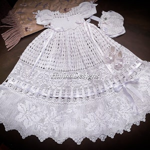 Puede incluir: Un vestido de bautizo blanco hecho a mano con gorro y patucos a juego. El vestido presenta un delicado diseño calado, detalles de lazo y un dobladillo festoneado. El conjunto se muestra sobre una superficie oscura. El texto "Halina Designs" es visible.
