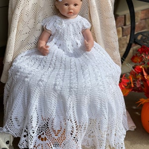 Puede incluir: Un vestido de bebé blanco de ganchillo con una diadema de flores. El vestido tiene un dobladillo festoneado y está hecho con encaje delicado. El vestido está sentado sobre una calabaza.