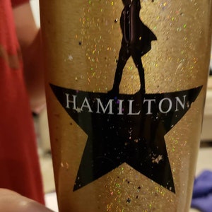 Op de afbeelding: Een gouden glitterbeker met een zwart silhouet van een persoon die op een zwarte ster staat met het woord "HAMILTON" erop.