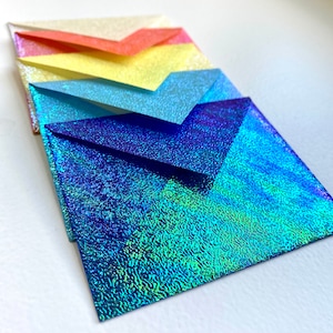 May include: A stack of five iridescent envelopes in a rainbow color scheme. The envelopes are arranged from top to bottom in the following order: white, pink, yellow, light blue, and blue.
