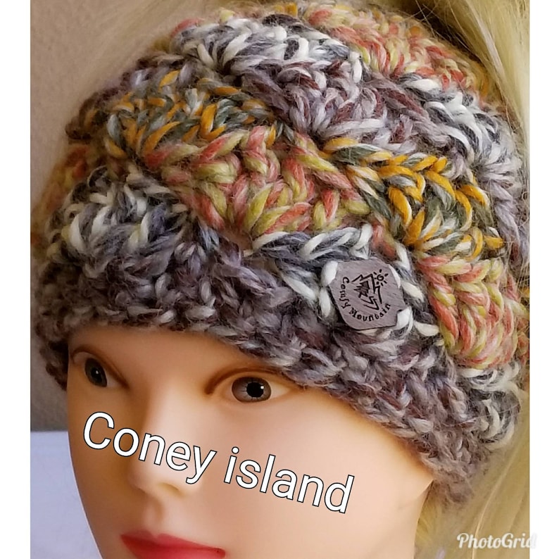 May include: A multi-colored crocheted headband with a brown leather tag that says "Crafty Mountain". The headband is made with a variety of colors, including gray, white, yellow, orange, and pink.
