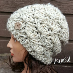 May include: A white crocheted beanie with a textured pattern and a small wooden tag that says "Snug Mountain". The beanie is shown on a mannequin with brown hair. The text "Oatmeal" is written in the bottom right corner of the image.