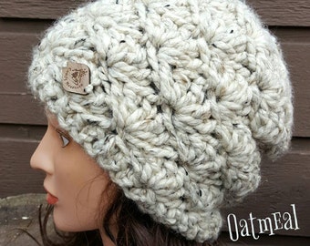 May include: A white crocheted beanie with a textured pattern and a small wooden tag that says "Snug Mountain". The beanie is shown on a mannequin with brown hair. The text "Oatmeal" is written in the bottom right corner of the image.