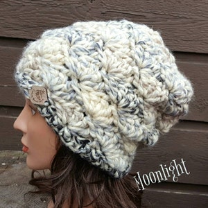 May include: A white, gray, and black crocheted beanie with a brown leather tag that says "Crafty Mountain". The beanie has a slouchy fit and is made with a chunky yarn.