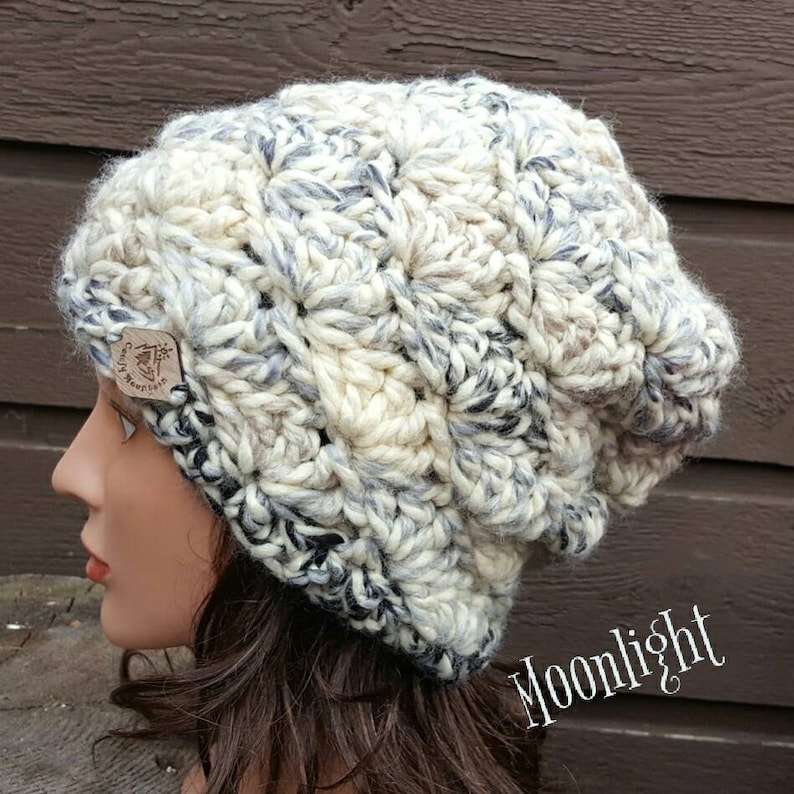 May include: A white, gray, and black crocheted beanie with a brown leather tag that says "Crafty Mountain". The beanie has a slouchy fit and is made with a chunky yarn.