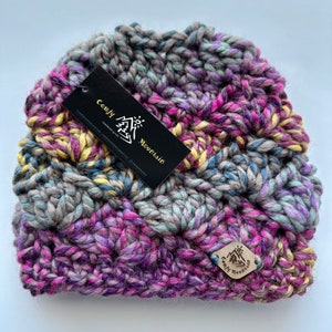 May include: A gray, purple, and yellow crocheted beanie with a brown leather tag that says "Cozy Mountain" in gold lettering.
