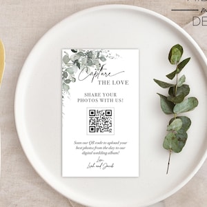 May include: A white card with a green leafy design and a black and white QR code. The text on the card reads "Capture the Love, Share Your Photos With Us!" and "Scan our QR code to upload your best photos from the day to our digital wedding album!"