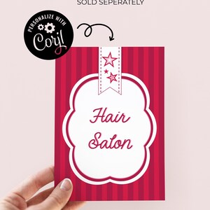 May include: A red and white sign for a hair salon, held by a hand. The sign has a white, cloud-shaped border with the words "Hair Salon" in cursive. The background is red with vertical stripes. The sign also has a star design.