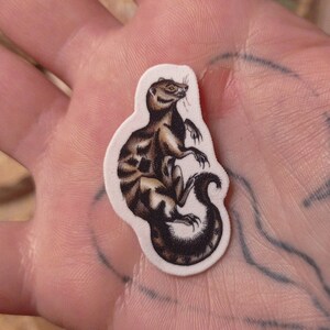 May include: A close-up of a small, white sticker with a black and brown illustration of a weasel-like creature. The sticker is being held in a person's hand.