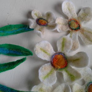 May include: Handmade felt flowers and leaves in a craft project. The flowers are white with orange and purple centers. The leaves are green with blue accents. The craft project is on a white background.