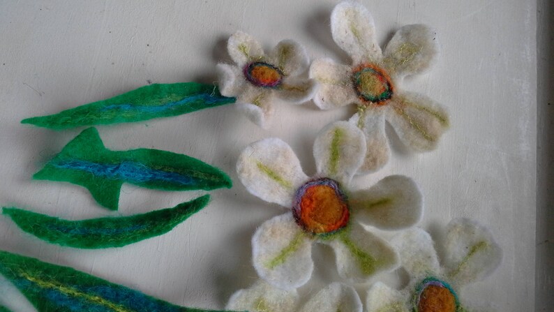 May include: Handmade felt flowers and leaves in a craft project. The flowers are white with orange and purple centers. The leaves are green with blue accents. The craft project is on a white background.
