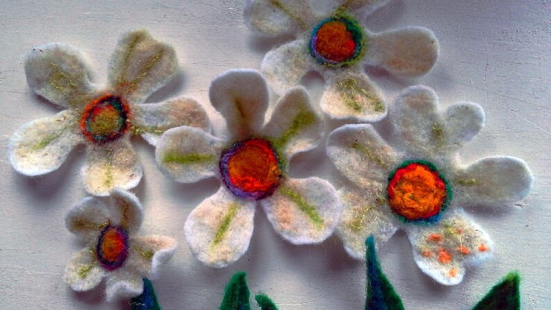 May include: Handmade felt flowers with white petals, gold accents, and vibrant orange, red, and blue centers. The flowers are arranged with green felt leaves, creating a whimsical floral design. Perfect for crafts or home decor.