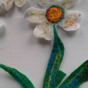 May include: Handmade felt flower art. The image shows a close-up of a felt flower with white petals, a yellow and orange center, and green and blue leaves. The flower is made of soft, textured felt.