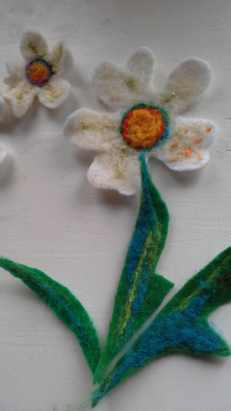 May include: Handmade felt flower art. The image shows a close-up of a felt flower with white petals, a yellow and orange center, and green and blue leaves. The flower is made of soft, textured felt.