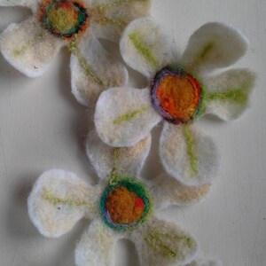 May include: Handmade felt flowers with white petals and colorful centers. The flower centers feature orange, green, and purple accents. The petals have green stitching details. These decorative flowers are perfect for crafts.