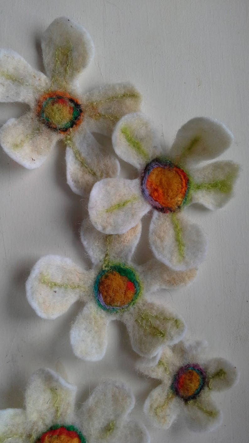 May include: Handmade felt flowers with white petals and colorful centers. The flower centers feature orange, green, and purple accents. The petals have green stitching details. These decorative flowers are perfect for crafts.