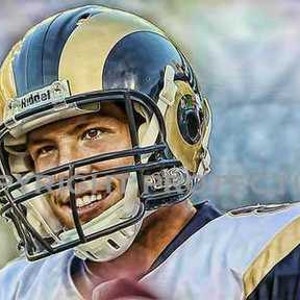 May include: A football player wearing a blue and gold helmet with a white face mask and a white and gold jersey. The player is smiling and looking to the right.