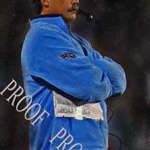 May include: A man wearing a blue jacket with a white logo on the left sleeve, brown trousers, and a headset with a microphone. He is looking to the right.